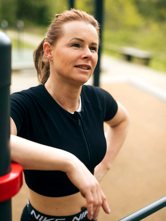 Halbkörper-Porträt von Deike im Freien, sie trägt ein schwarzes Top, schaut nach rechts oben aus dem Bild und lehnt ihren rechten Arm locker auf.